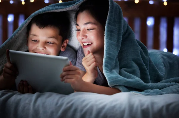 Two kids holding digital tablet while lying under blanket in the dark at night reading online book, watching or playing game before sleeping. Little boy and sister lying in bed and faces illuminated