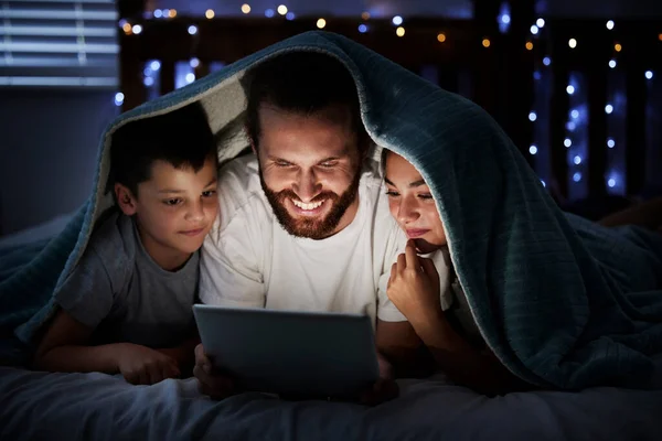 Happy caucasian family single dad with two children using digital tablet lying under blanket in the dark at night with their faces illuminated by device screen light. Father reading online story or