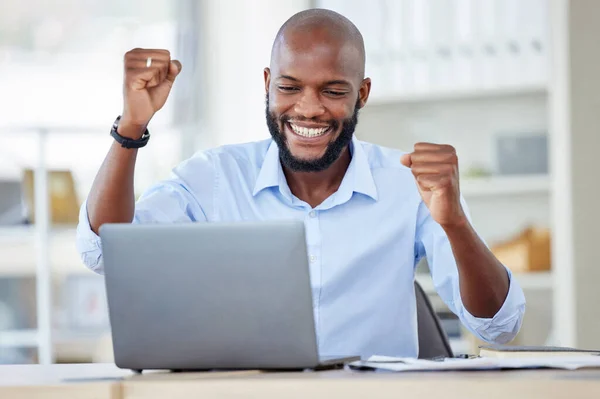 Young happy african american businessman cheering with joy and working on a laptop in an office at work alone. One cheerful male business professional cheering with his fists and using a computer