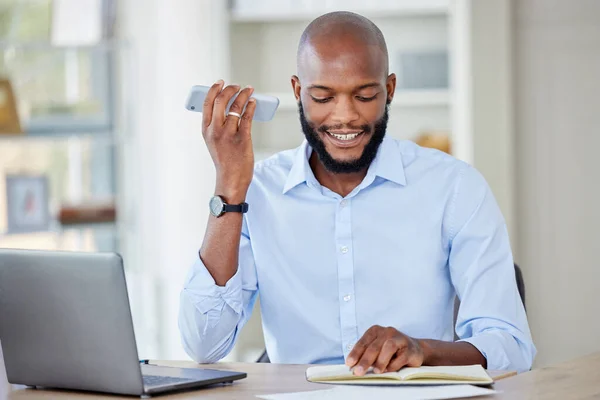 Young african american businessman on a call using a phone while reading a note in a notebook and working on a laptop in an office at work alone. One male business professional talking on a cellphone