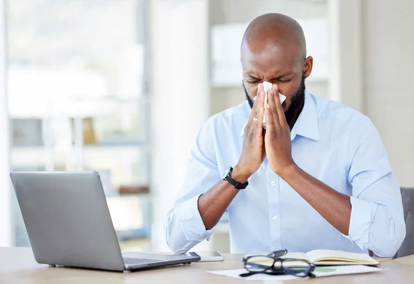 Young african american businessman blowing his nose with a tissue while sitting at a desk at work. Sick male businessperson using a laptop while suffering from allergies at work.