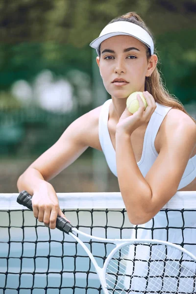 Dedicated young female tennis player holding a tennis racket and ball while leaning over a net. Hispanic woman ready for her tennis match at the club. Sportswoman ready for tennis practice.