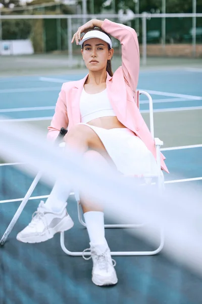 Trendy young tennis player wearing a pink jacket while sitting on chair on a tennis court. Young hispanic sportswoman sitting with a tennis racket after a match.