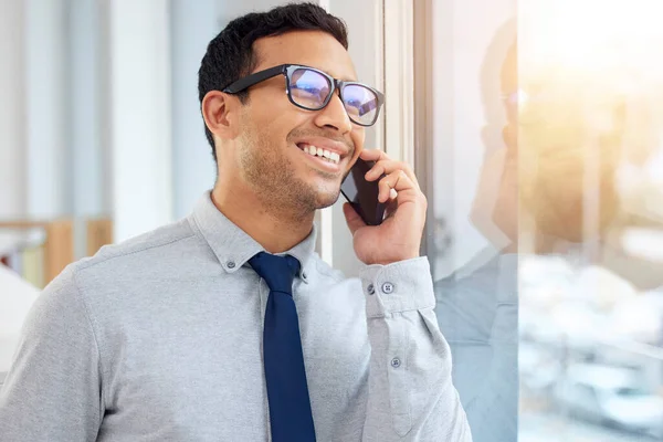 Young content mixed race businessman on a call using a phone and standing in an office at work. One hispanic male businessperson smiling while talking on the phone standing and thinking at a window in