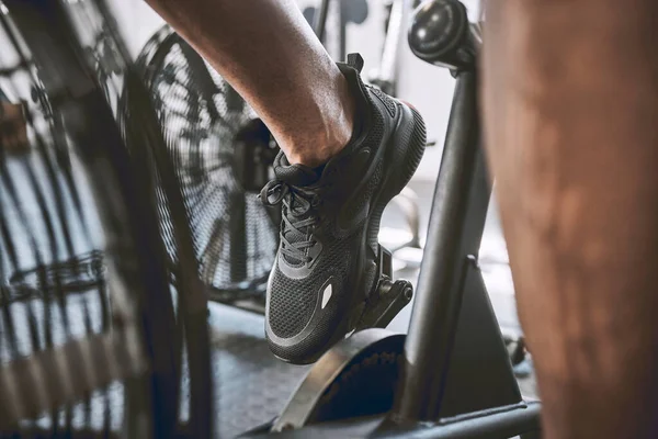 Foot of a fit man cycling in the gym. Trainer cycling in sport shoes in the gym. Bodybuilder riding a gym bike cropped. Wear the right shoes when exercising at the gym. Building strong legs on a bike.