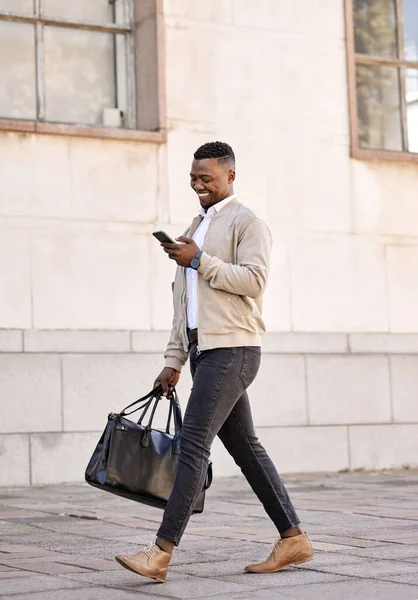 Black businessman travelling alone.A african american businessman walking around town with his luggage while looking at his smartphone and smiling in the city.