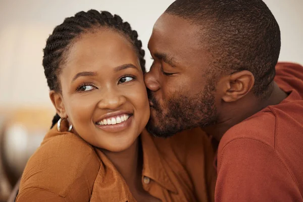 Loving young african american man kissing his wife on the cheek while she smiles and looks away. Happy young man and woman sharing romantic intimate moment at home.