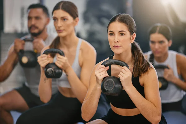 Active young caucasian woman doing kettlebell squat exercises while training together with a group of people in a gym. Focused athletes challenging themselves by holding heavy weights to build muscle