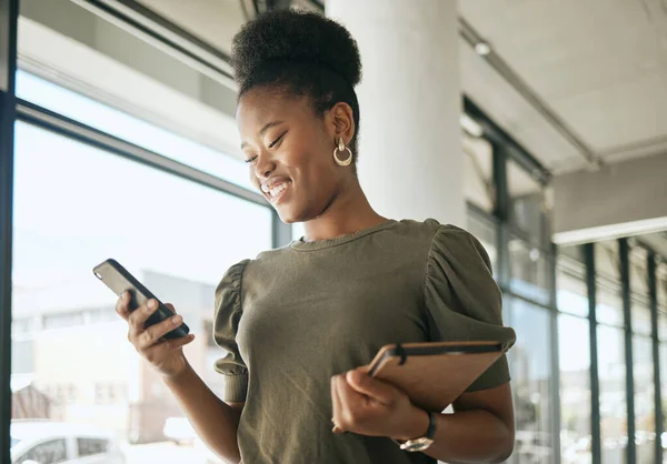 African american business woman smiling while reading or texting on smartphone holding journal and walking in modern office. Smiling female entrepreneur using mobile app or browsing social media.