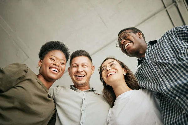 Team of happy united young business people hugging each other. Diverse mixed race group of men and women standing in row in their office, huddling and laughing together.