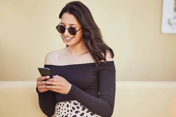 Fashionable young mixed race woman with sunglasses using her smartphone while standing on sidewalk. Millennial woman sending a text or chatting on social media while on city street.