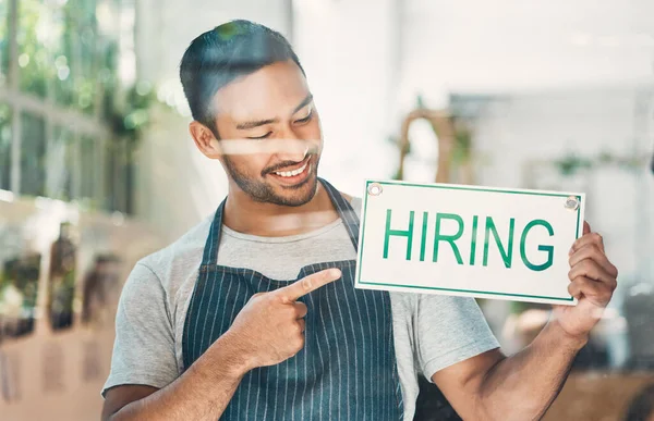 One young hispanic man pointing to a hiring sign at a window on display in a cafe or store. Happy mixed race guy advertising job opportunity while recruiting new staff for his startup shop.