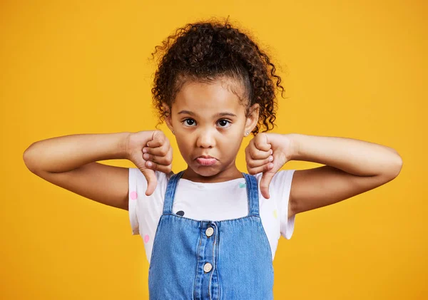 Studio portrait mixed race girl giving thumbs .down isolated against a yellow background. Cute hispanic child posing inside. Unhappy and upset kid being negative and saying, I disagree or disapprove.