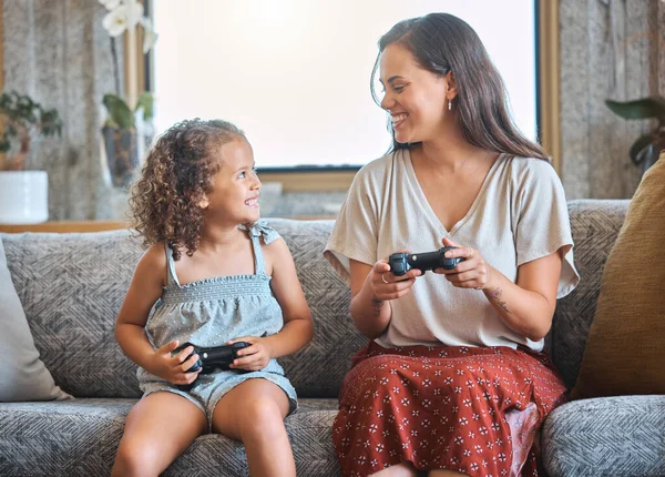 Hispanic mother and daughter playing video games together while sitting on the couch at home. Fun young mother and daughter using joysticks while playing and spending free time together on weekend.