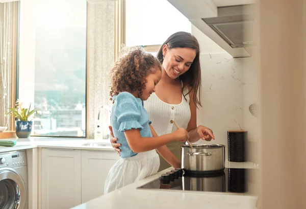 Mother and little daughter preparing dinner together at home. Young mother standing with her daughter and helping her while stirring food on the stove. Mom teaching little girl to cook.