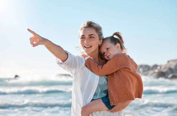 Mother and daughter on vacation by the sea. Child enjoying a getaway with her caucasian mom on a bright summer day, smiling family relaxed against a bright copyspace background while pointing.