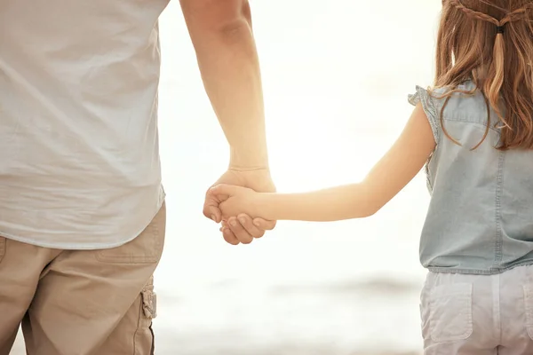 Back of a father and daughter holding hands on the beach at sunset. Father bonding with his child, being affectionate and keeping her safe by holding her hand outside. Parent and child together.