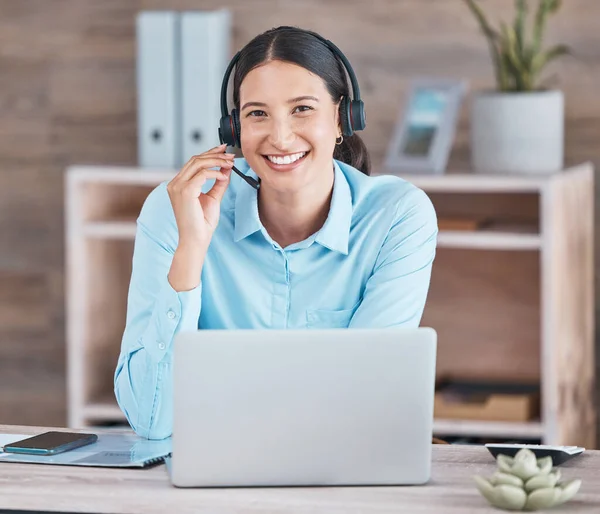 Portrait of a happy smiling hispanic woman wearing a headset and using a laptop while sitting at a desk at her office job. Cheerful young mixed race woman looking positive while working online.