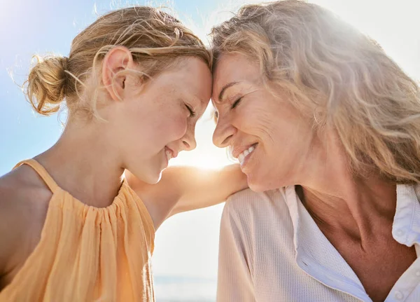 Cheerful mature woman and little girl talking and sharing a secret while sitting on the beach. Happy little girl smiling while sitting with her mom or grandmother and being loving and affectionate.