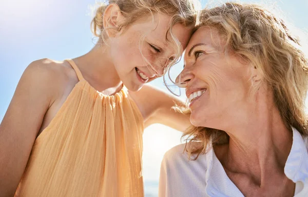 Cheerful mature woman and little girl talking and sharing a secret while sitting on the beach. Happy little girl smiling while sitting with her mom or grandmother and being loving and affectionate.