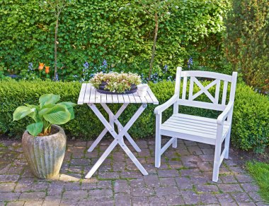 White wooden chair and table in a green garden with blossoming plants and copyspace. Tranquil landscape to relax and enjoy a cosy picnic in summertime. Outdoor patio in a quiet and secluded courtyard. clipart