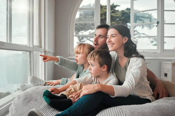 Young caucasian couple enjoying the view from a window while bonding with their adorable little children on a comfy seat. Cute kid looking amazed and pointing in surprise while sitting with her family