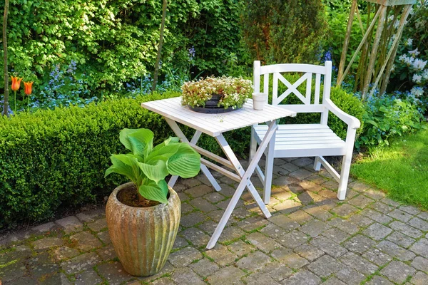 Patio chair and square table with lots of plants in a garden. Outdoor furniture for relaxing in summer while flower gardening. Potted plants in a small paved courtyard with green grass and trees.