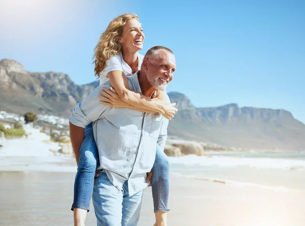 Happy mature couple enjoying vacation by the beach. Active senior husband giving his wife a piggyback ride while enjoying a sunny day outdoors. Energetic man and woman having fun while on holiday.