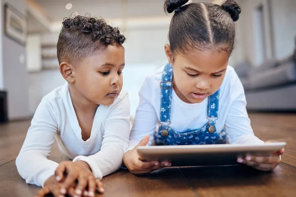 Two young mixed race children using a digital tablet while lying together on the floor at home. Young boy and girl siblings browsing the internet online to play games and learn from educational apps.