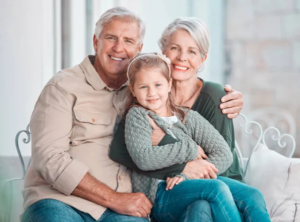 Little girl and her grandparents looking at the camera while sitting together at home. Cute girl bonding with her grandmother and grandfather. Loving couple spending time with their granddaughter.