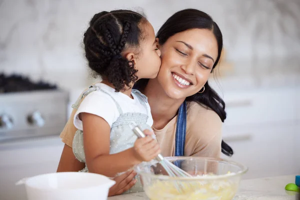 Happy latin mother and daughter baking and sharing a kiss while bonding. A young woman helping her daughter bake or cook while stirring the batter and giving an affectionate hug in the kitchen.