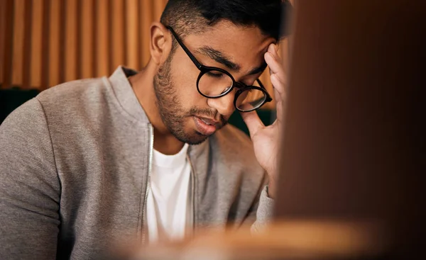 Young stressed indian man or student struggling with headache while browsing on a laptop in a cafe. One tired and frustrated guy suffering with burnout and pressure while studying or working freelance