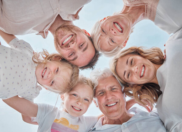 Below shot of multi-generation family smiling in a huddle against the blue sky. Carefree family with two children, parents and grandparents standing together and looking down at the camera.