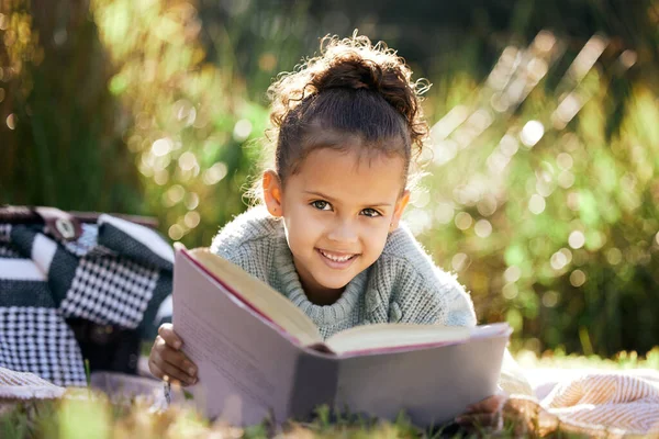 A little girl reading a book while relaxing in a park or garden. Mixed race child learning and getting an education, lying on the grass in nature and having fun.