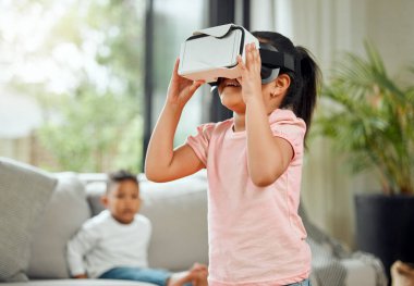 a little girl playing with a virtual reality headset while her brother watches from the sofa at home.