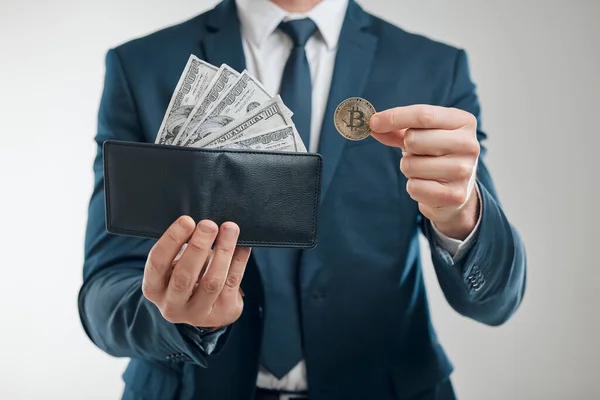 Closeup shot of an unrecognisable businessman holding a bitcoin and a wallet filled with banknotes against a white background.