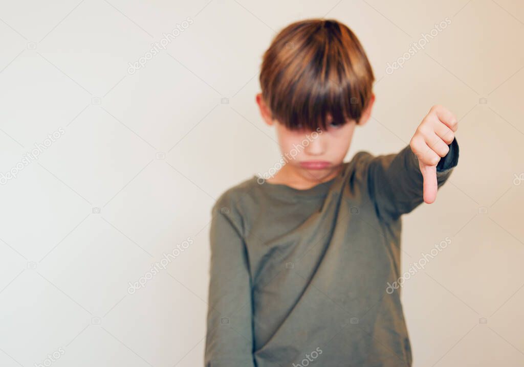 Studio shot of a cute little boy showing the thumbs down against a wall.