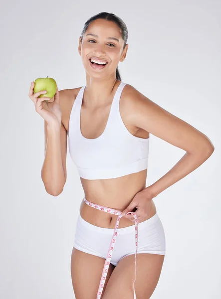 Studio portrait of an attractive young woman eating an apple and using a measuring tape against a grey background.