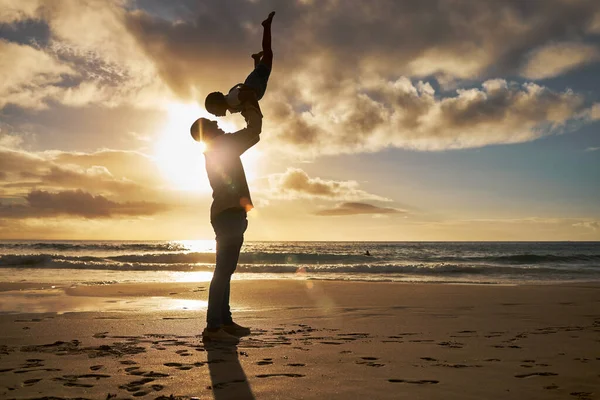 Little girl playing and bonding with her dad on vacation. Silhouette of a loving father holding up his little child on the beach against golden sky. Parent lifting his daughter against sunset sky