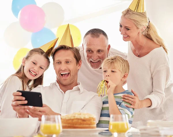 A happy family celebrating a birthday with a party, wearing hats and taking a selfie using a phone. Mature man taking a photo of his father, wife and children at a party while making special memories.