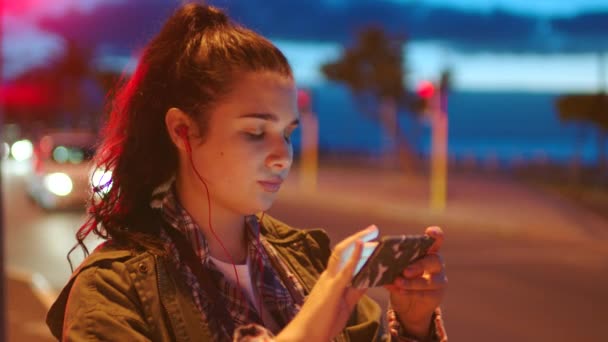Poważna Młoda Kobieta Używająca Telefonu Nosząca Słuchawki Stojąca Nocą Ulicy — Wideo stockowe