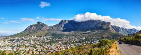 A mountain road overlooking the city with a cloudy blue sky. Panoramic landscape of green mountains surrounding an urban town and a scenic road for traveling along Cape Town, South Africa.
