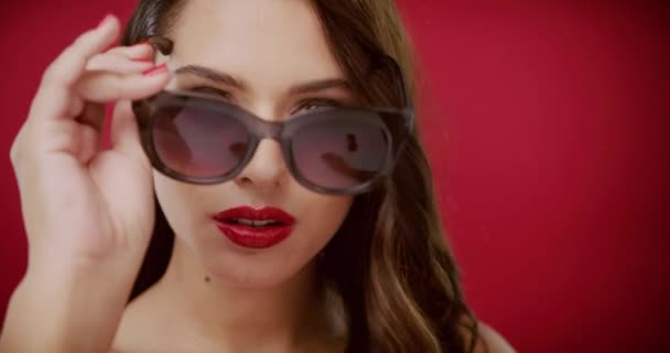 Відеозапис Красивої Молодої Жінки Надягає Сонцезахисні Окуляри Червоний Студійний Фон — стокове відео