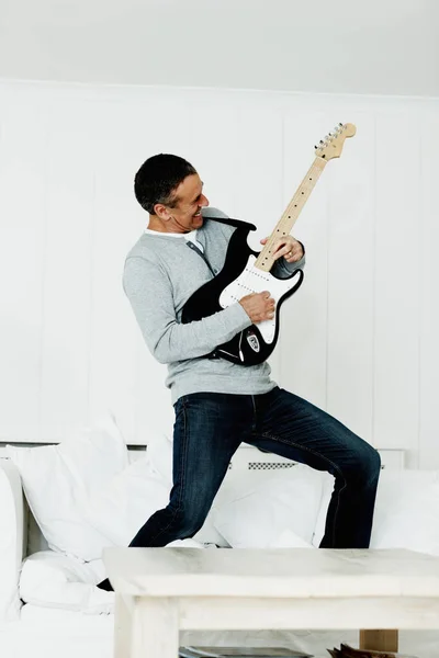 Portrait of an energetic mature man standing on sofa playing an acoustic guitar.