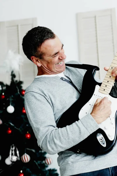 Portrait of an energetic mature man playing guitar with Christmas tree in background.