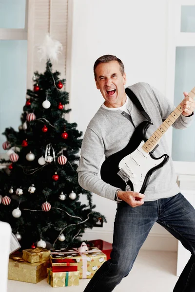 Portrait of a cheerful mature man playing guitar with Christmas tree in background.