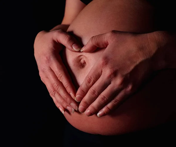 Detail shot of a pregnant woman making heart shape with hands against black background.