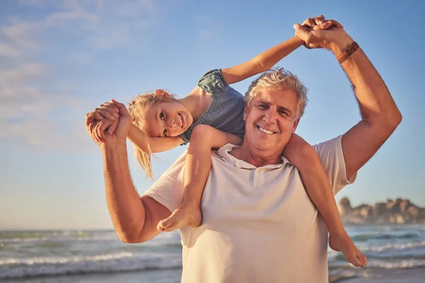 Portrait of Grandfather carrying his granddaughter on his shoulders while walking along the beach. Adorable little girl sitting on grandpas shoulders while holding hands and smiling.