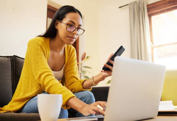 Woman working remote while typing on her laptop and holding her smartphone sitting on a sofa in a bright living room. One focused young hispanic female with glasses at home using modern technology.