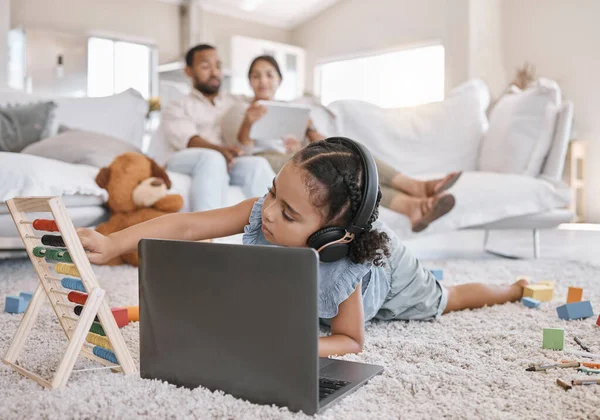 Closeup of a little cute girl using a laptop and wireless headphones while laying on the floor in the lounge. Hispanic girl using a wireless device to do her homework in the living room.